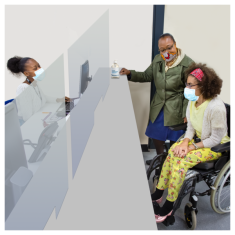 Three people pictured. One person  is on a computer behind a grey screen. On the other side of the screen are the other two people. One of these people is sitting in a wheel chair and the other is standing up slightly facing the person in the wheelchair.  