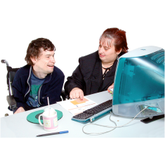 two people sitting at a table behind a computer. One person is pointing to a piece of paper on the table. 