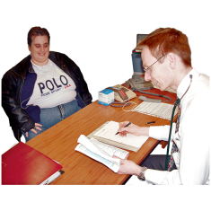 A doctor is sitting across a table from a person. The doctor is holding a book in his left hand and writing on a note pad with his other hand. 