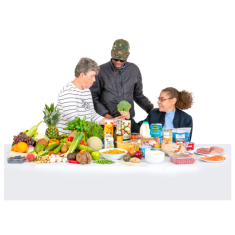 A table filled with healthy foods like meats, fish, vegetables and fruits. Behind the table are three people looking and talking about the food. 