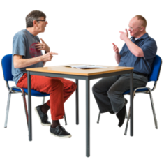 Two people sitting at a table. They are facing each other and communicating using sign language. 